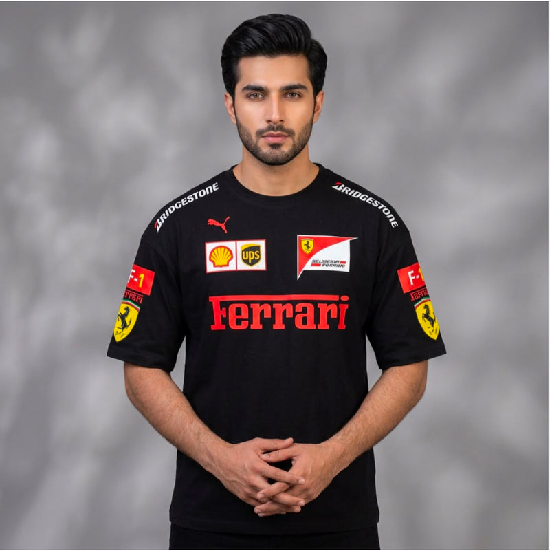 A man wearing a black Ferrari short-sleeve sports jersey with red and yellow sponsor logos, including Bridgestone, Puma, Shell, and UPS, standing against a gray studio background.